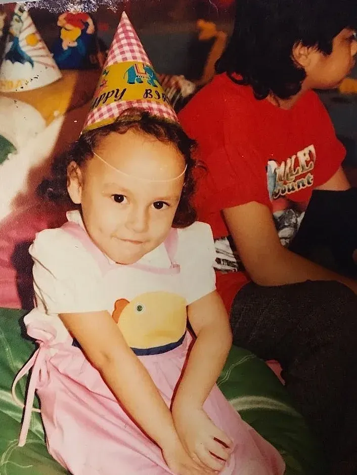 Snapshot of a young child in a party hat being embraced at a birthday celebration, with a colorful banner in the background.