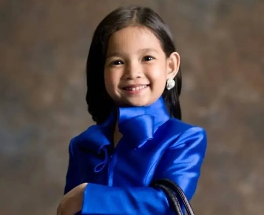 Studio portrait of a smiling young girl with dark hair wearing a shiny royal-blue long-sleeved outfit, posing with arms folded against a brown backdrop.