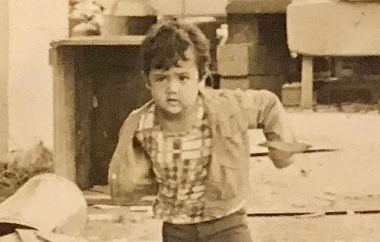 Sepia-toned vintage photo of a young boy with curly hair in a checkered shirt, captured mid-motion reaching forward in what appears to be a film scene.