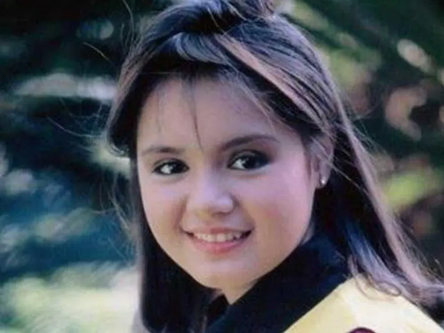 Vintage portrait of a smiling young girl with long dark hair and side-swept bangs, wearing pearl earrings and a yellow-and-black top, photographed outdoors against blurred greenery.