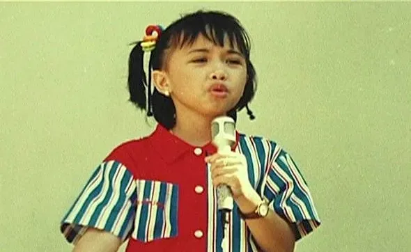 Vintage photo of a young girl with pigtails wearing a striped shirt, singing into a microphone against a green background.