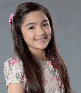 Portrait of a smiling young girl with long brown hair and a flower hairclip, wearing a floral top against a plain background.