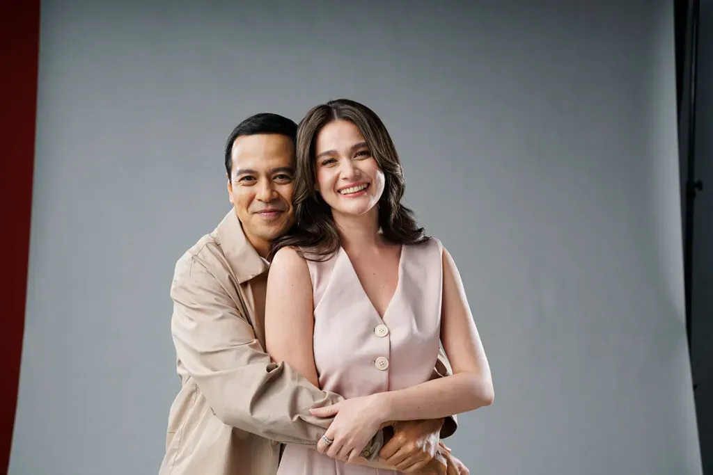 Photo of a smiling couple in coordinated neutral outfits posed close together against a plain background.