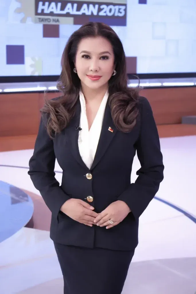 Portrait of a woman in a dark suit standing in a television news studio with an “HALALAN 2013” screen behind her.