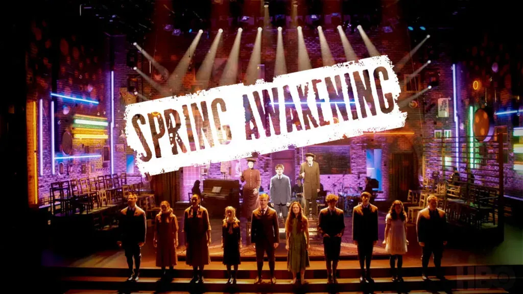 Stage photograph of a theatrical production with performers on a brightly lit set beneath a marquee-style sign reading “SPRING AWAKENING.”