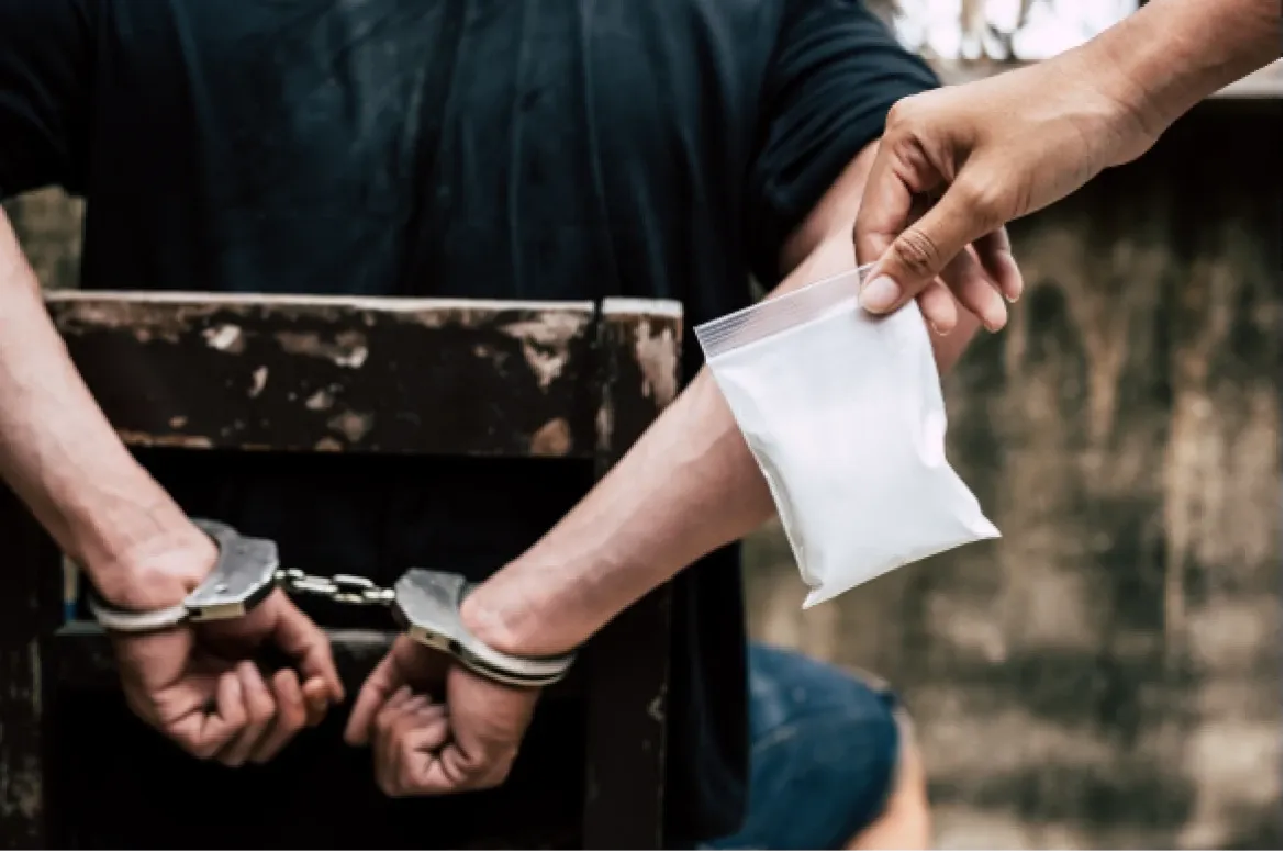 Editorial photo of a person’s handcuffed wrists beside another hand holding a small plastic sachet, representing an anti-drug operation.
