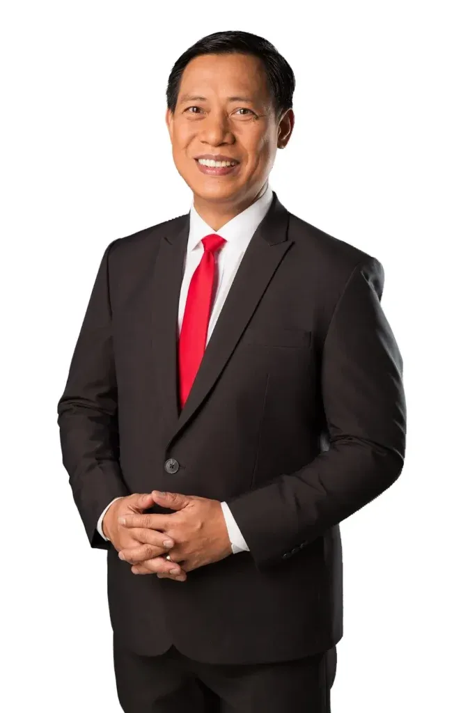 Studio portrait of a man in a dark suit with a red tie, standing with hands clasped against a white background.