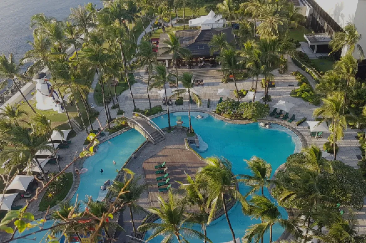 Photo of a resort swimming pool surrounded by palm trees and lounge areas, viewed from above.