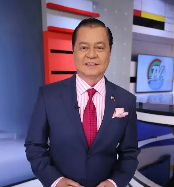 Portrait of a man in a blue suit and red tie smiling in a television studio with screens behind him.