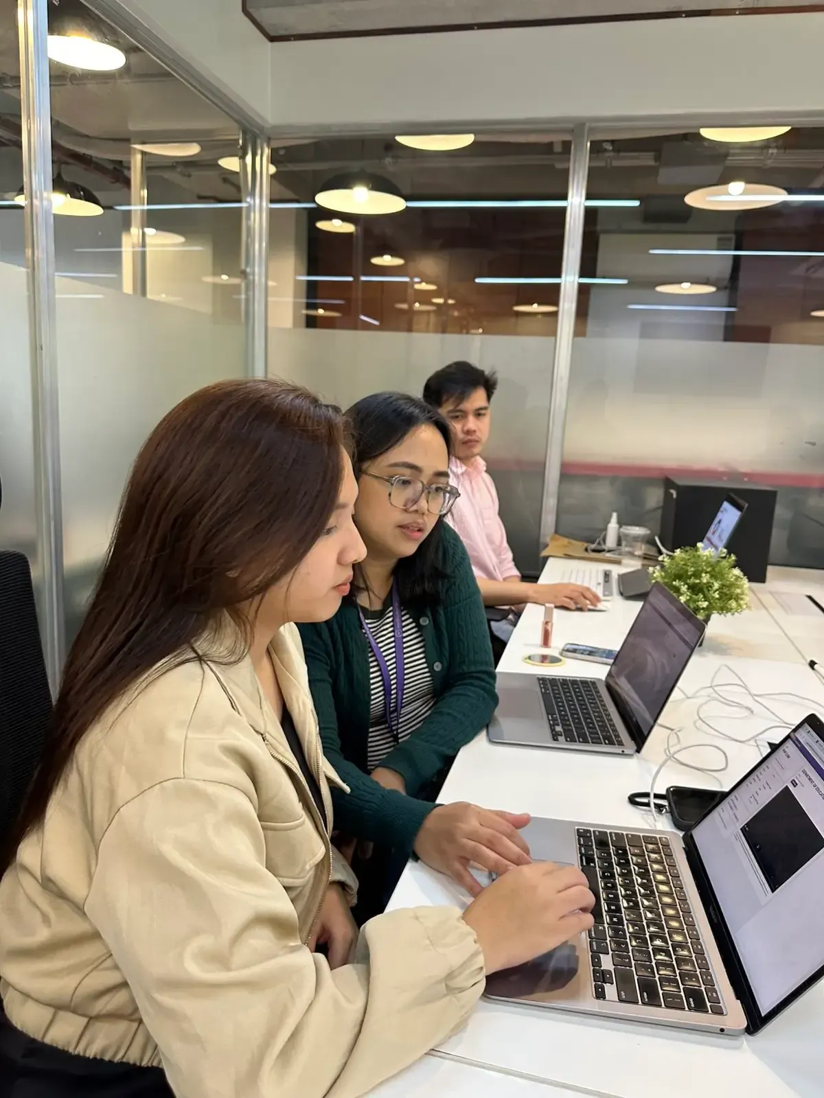 Photo of two colleagues working together on a laptop at a table in a bright modern office, with others in the background.