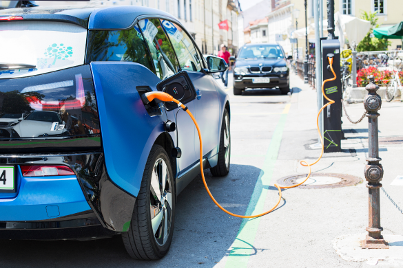 Photo of a blue electric car plugged into a charging station on a city street.