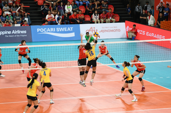 Photo of a women’s volleyball match in progress, players in yellow jerseys at the net with advertising boards behind.