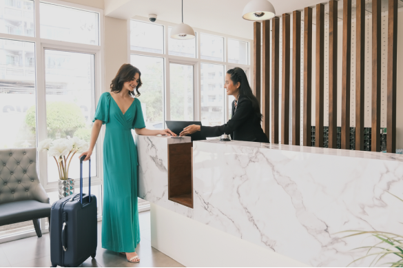 Photo of a hotel reception desk where a staff member in a teal uniform assists a guest with luggage in a marble lobby.