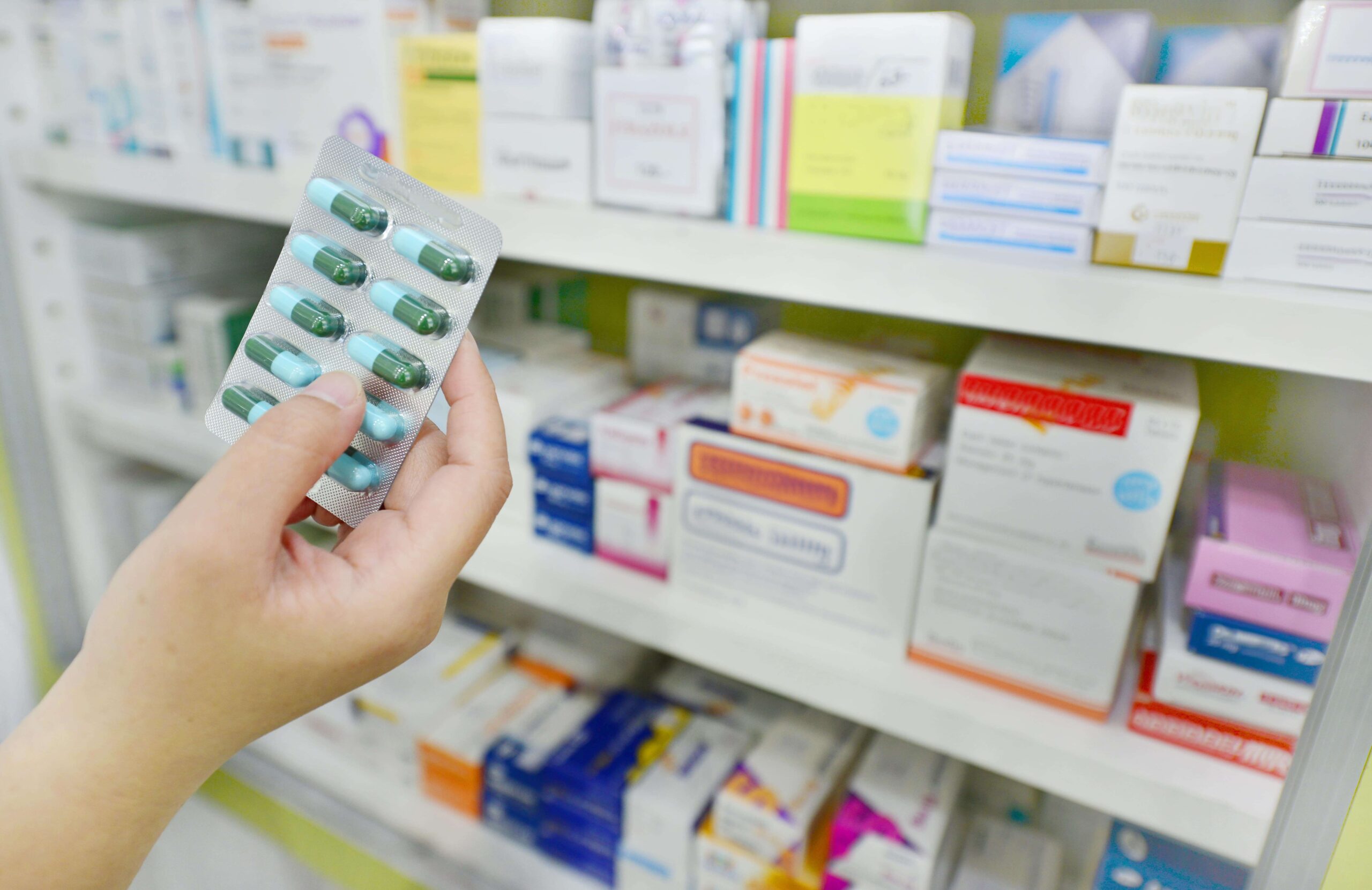Photo of a hand reaching for blister packs of medicine on a pharmacy shelf stocked with boxes.